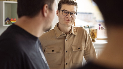 Billede af en ung mand med briller og et venligt smil, iført en beige skjorte med lommer, der står i et lyst og indbydende rum, mens han interagerer med andre personer i en uformel samtale.